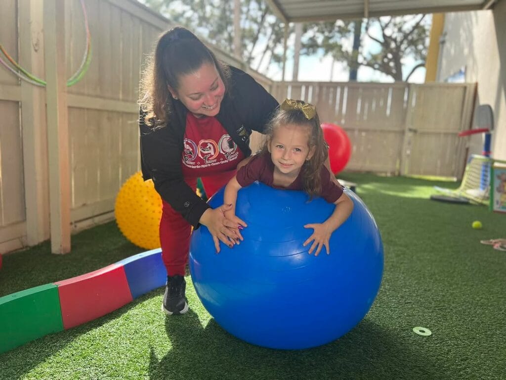 Physical therapist providing outdoor balance and coordination therapy with young girl using large blue therapy ball
